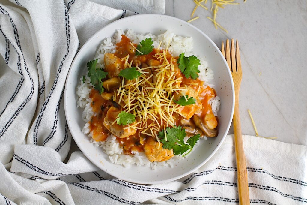 Brazilian Chicken Stroganoff in a tomato-based sauce in a bowl over white rice with cilantro garnish and thin potato sticks on top. This is an easy 30 minute dinner with a tomato based sauce, cream, and mushrooms all over rice.