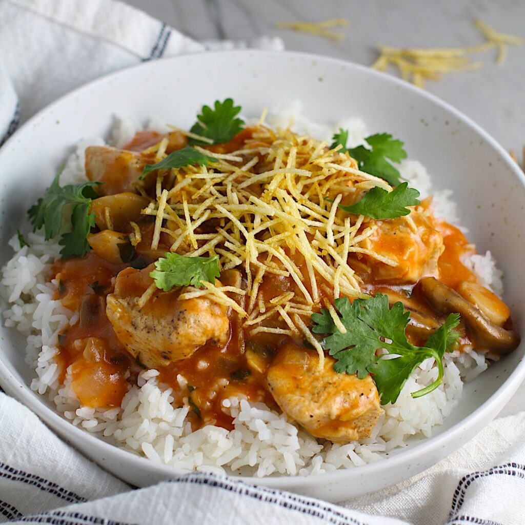 Brazilian Chicken Stroganoff in a tomato-based sauce in a bowl over white rice with cilantro garnish and thin potato sticks on top. This is an easy 30 minute dinner with a tomato based sauce, cream, and mushrooms all over rice.