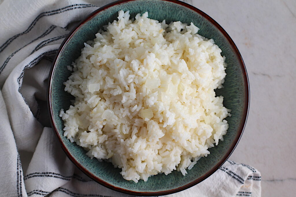Brazilian Rice in a bowl on counter next to a towel. It's fluffy white rice flavored with sauteed garlic and onion. It takes only a couple additional steps and goes with anything!