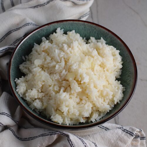 Brazilian Rice in a bowl on counter next to a towel. It's fluffy white rice flavored with sauteed garlic and onion. It takes only a couple additional steps and goes with anything!