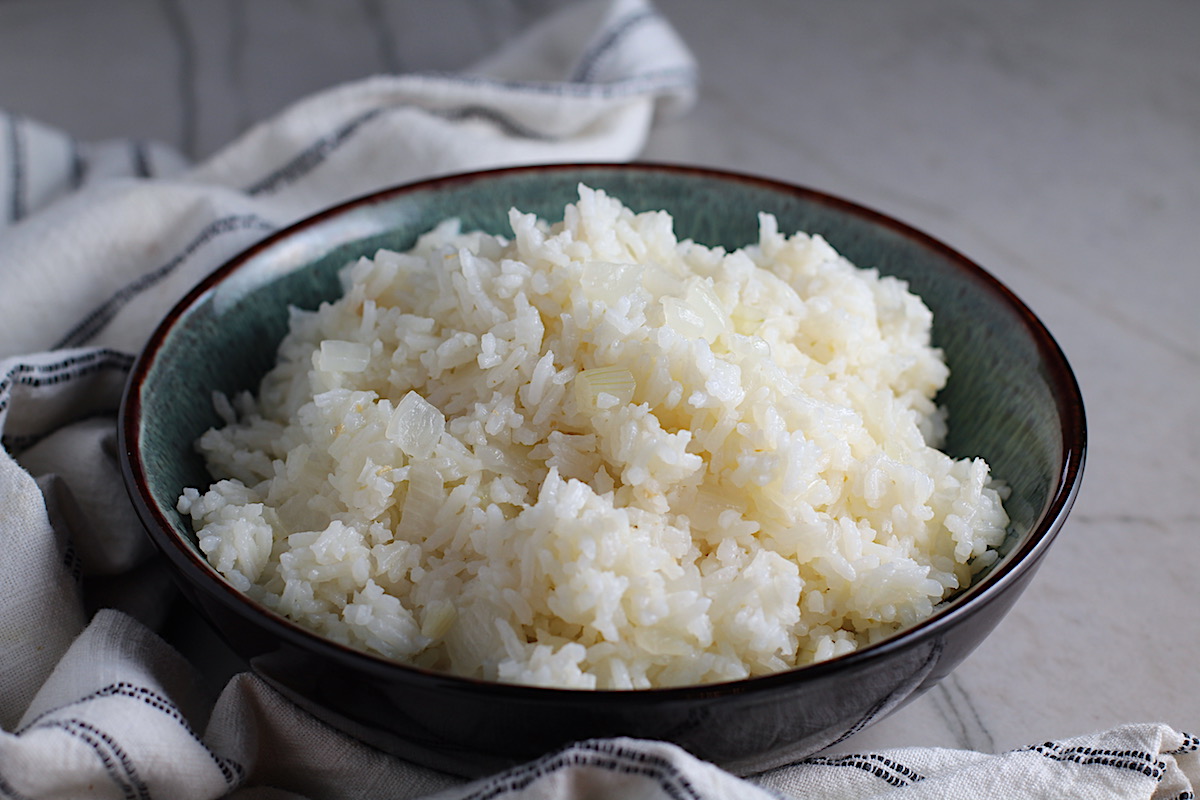 Brazilian Rice in a bowl on counter next to a towel. It's fluffy white rice flavored with sauteed garlic and onion. It takes only a couple additional steps and goes with anything!