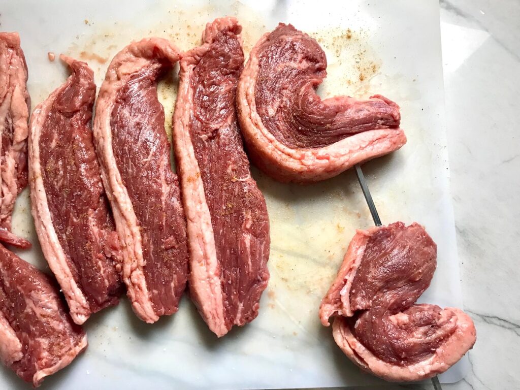 Slices of Picanha steak on a cutting board with 2 folded over being skewered for the Brazilian Steak recipe.