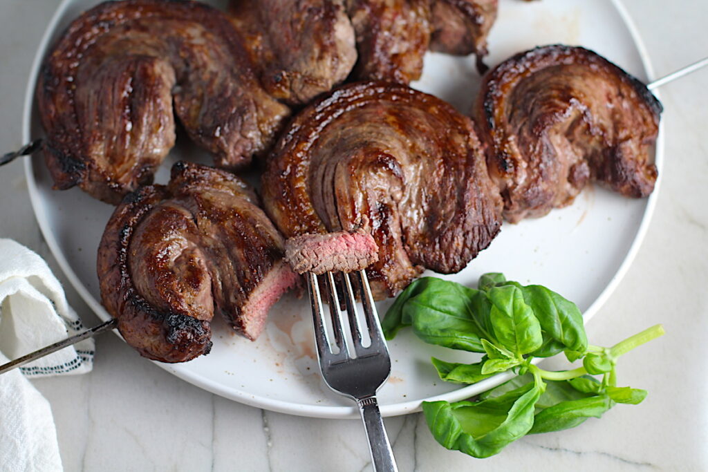 Picanha steak skewered on a plate with basil leaves and a fork holding a bite. It's the best Brazilian steak recipe, a tender and delicious cut of beef seasoned with a Brazilian blend and grilled perfectly!
