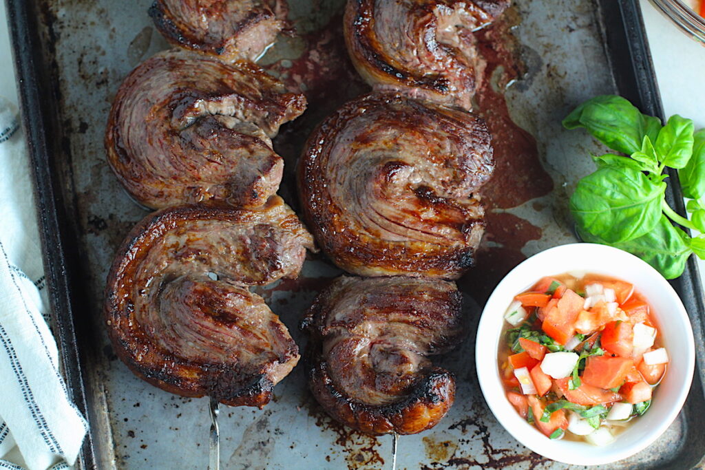 Picanha steak skewered on a sheet pan with a tomato salad and basil leaves. It's the best Brazilian steak recipe, a tender and delicious cut of beef seasoned with a Brazilian blend and grilled perfectly!