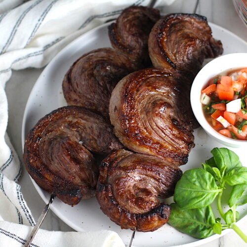 Picanha steak skewered on a plate with a tomato salad and basil leaves. It's the best Brazilian steak recipe, a tender and delicious cut of beef seasoned with a Brazilian blend and grilled perfectly!