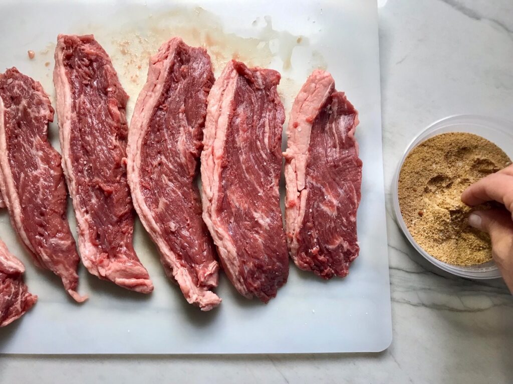 Hand pinching up some Brazilian Steak Seasoning from a small bowl to sprinkle on the raw steaks on the cutting board to the left.