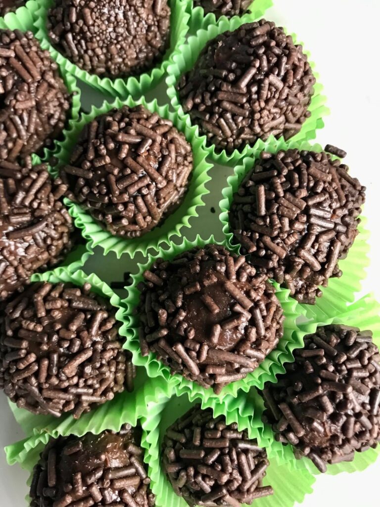 Brigadeiros in green paper in a plastic container. This Brigadeiro Recipe is based on traditional Brazilian chocolate truffles rolled in sprinkles. You won't believe how easy they are to make!