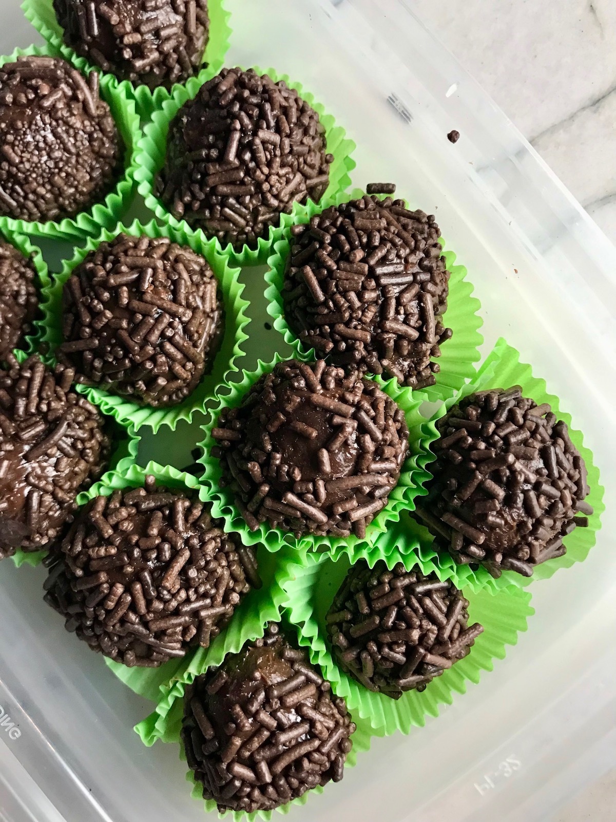 Brigadeiros, Brazilian Chocolate Dessert, in green paper in a plastic container. This Brigadeiro Recipe is based on traditional Brazilian chocolate truffles rolled in sprinkles. You won't believe how easy they are to make!