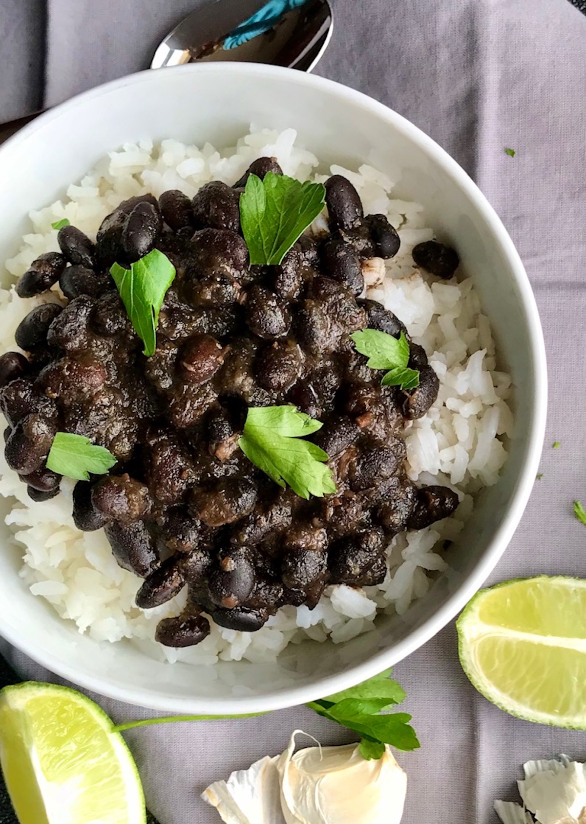 Brazilian Black Beans Recipe (Feijao) over rice in a bowl with cilantro leaves on top, lime wedges and garlic cloves on the side. It's delicious, thick, & hearty.