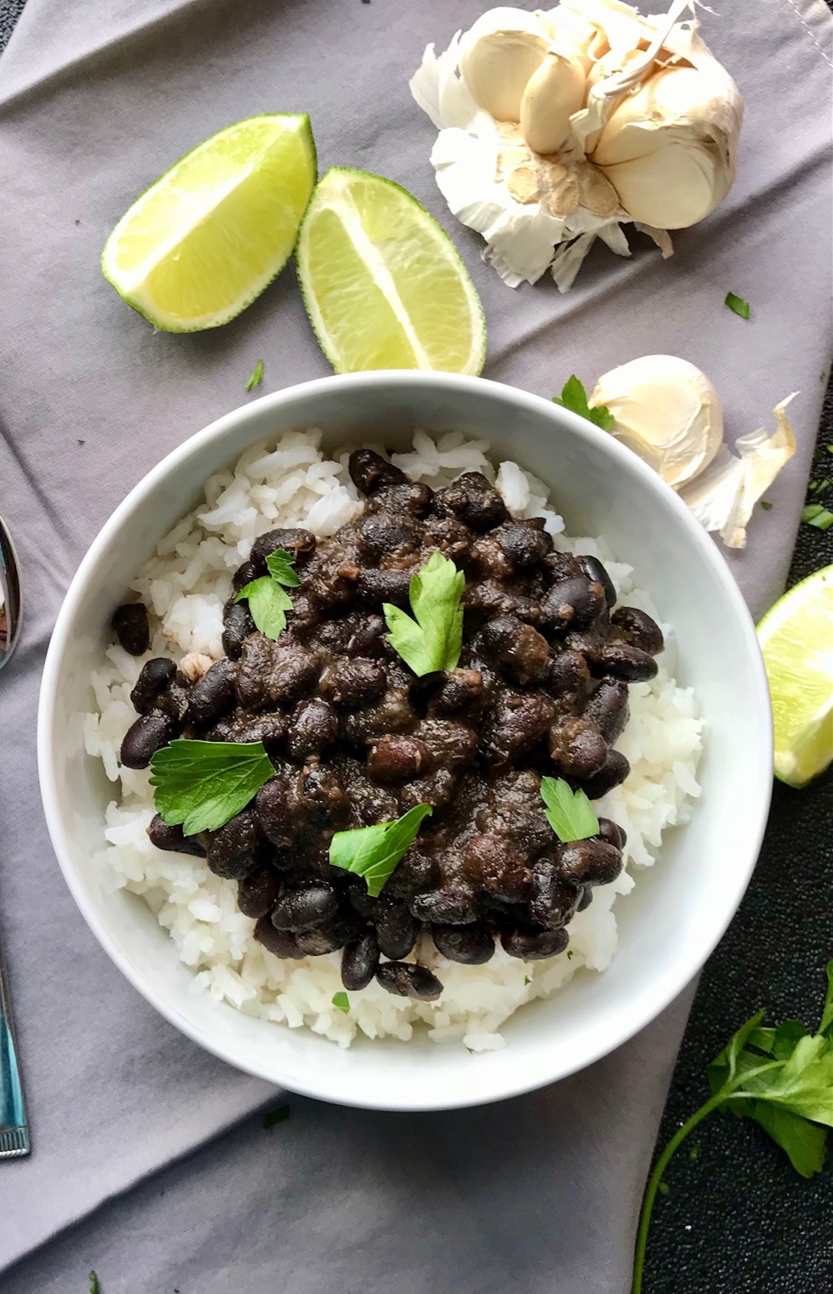 Brazilian Black Beans Recipe (Feijao) over rice in a bowl with cilantro leaves on top, lime wedges and garlic cloves on the side. It's delicious, thick, & hearty.