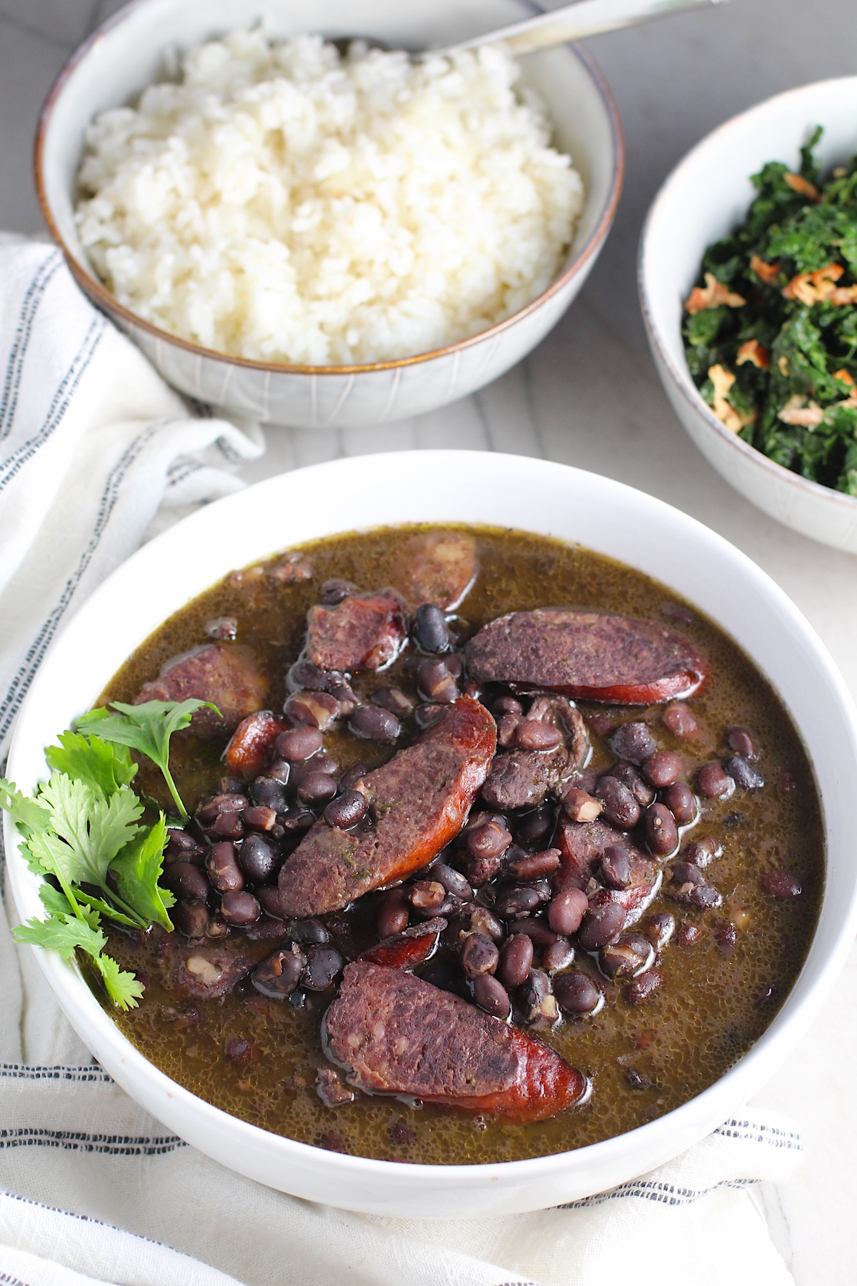 Bowl of cooked black beans with pork sausage slices and beef and cilantro garnish on side.  It is on table with a bowl of cooked kale and a bowl of rice with towel next to it.  Feijoada is rice and beans Brazilian style with pork sausages, beef, and more. 