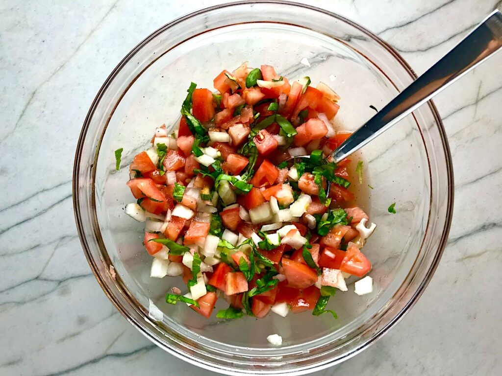 Tomato and onion salad in clear bowl on counter. It has diced tomato, diced onion, basil, and vinegar.