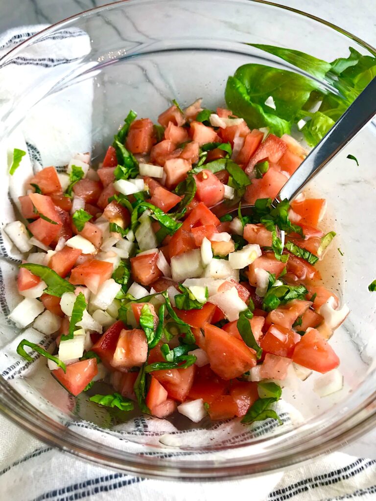 Tomato and onion salad in clear bowl on counter with towel. It has diced tomato, diced onion, basil, and vinegar.