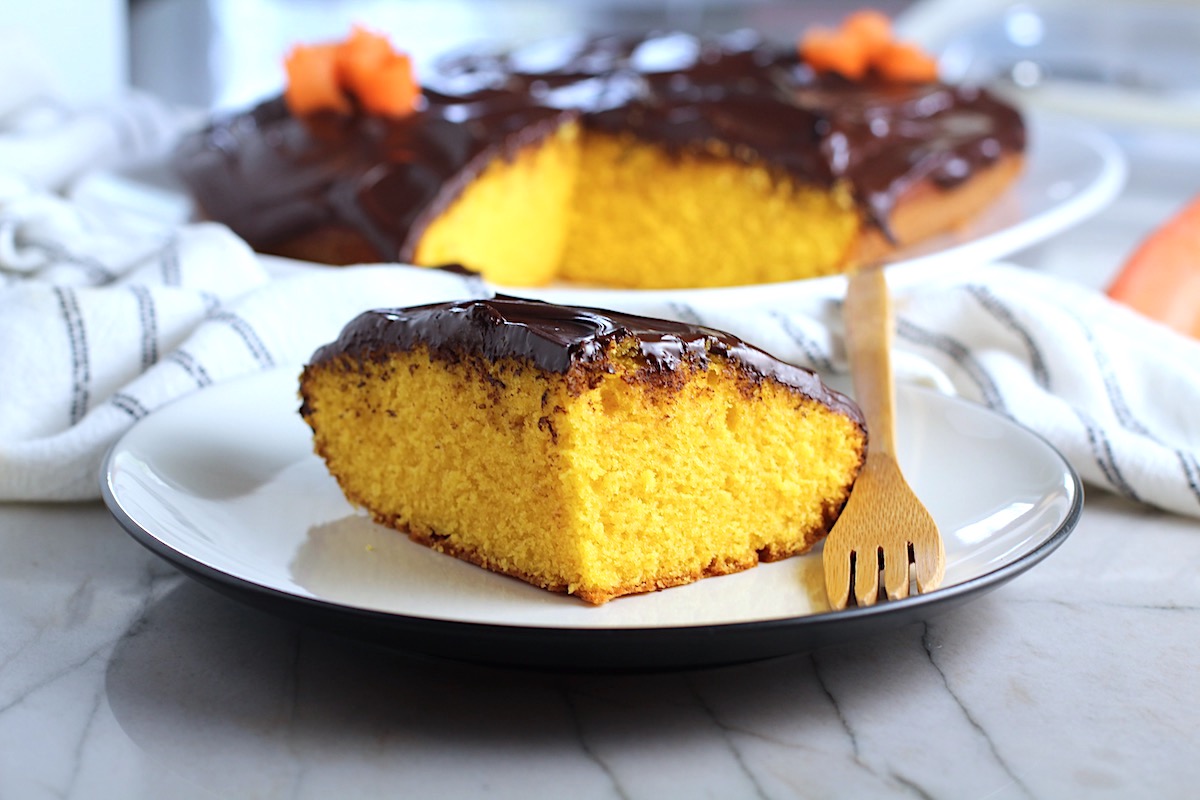 Slice of Brazilian Carrot Cake with Chocolate Sauce topping on a plate with bamboo fork and whole cake in background. It's absolutely delicious and easy to make!
