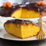 Slice of Brazilian Carrot Cake with Chocolate Sauce topping on a plate with bamboo fork and whole cake in background. It's absolutely delicious and easy to make!