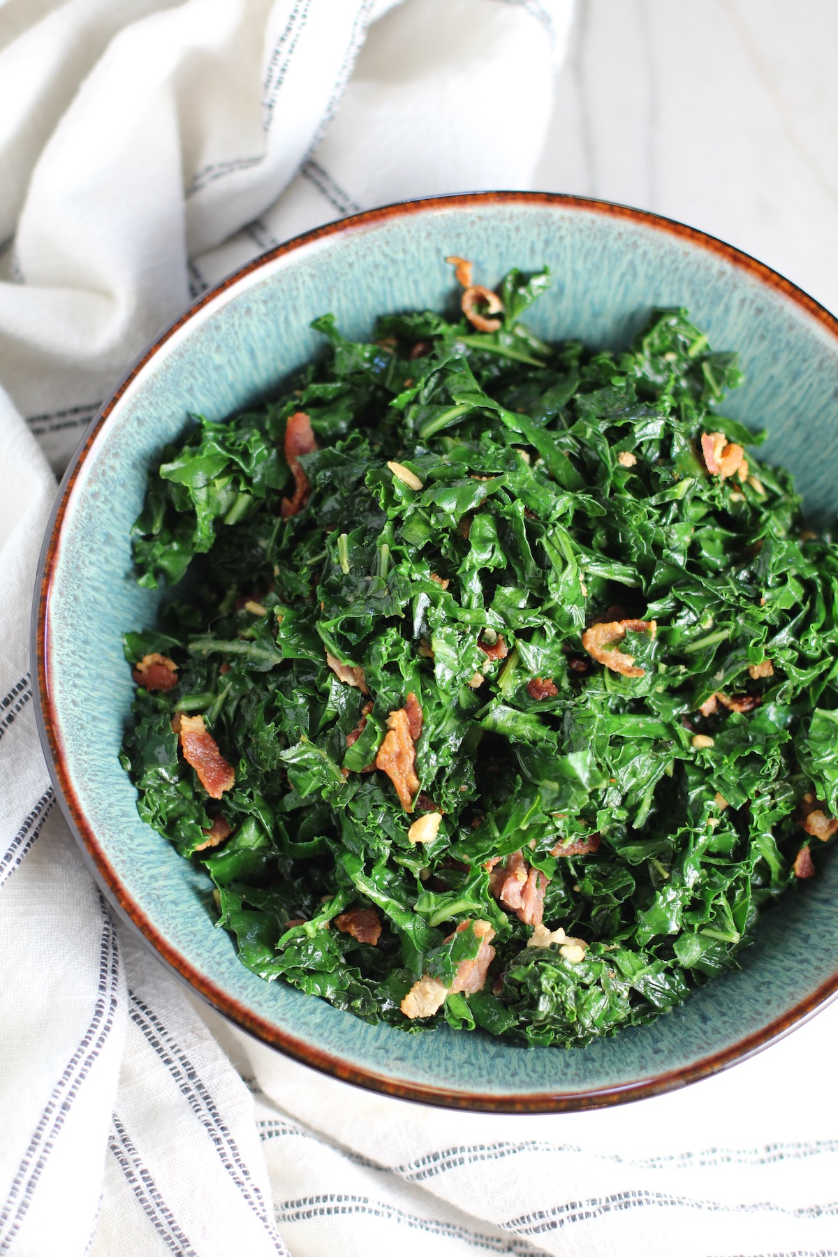 Brazilian sauteed Kale with Bacon and Garlic in a bowl on counter with white and blue towel. It's known as Couve Mineira in Portuguese, and is an easy and delicious side for any dinner!