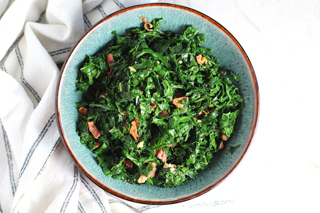 Brazilian sauteed Kale with Bacon and Garlic in a bowl on counter with white and blue towel. It's known as Couve Mineira in Portuguese, and is an easy and delicious side for any dinner!