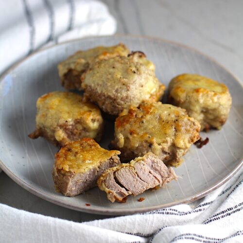 Parmesan Pork Tenderloin pieces on a plate with one cut in half. They are juicy, salty, and slightly cheesy. It's easy to make in the oven so you can enjoy it at home anytime!