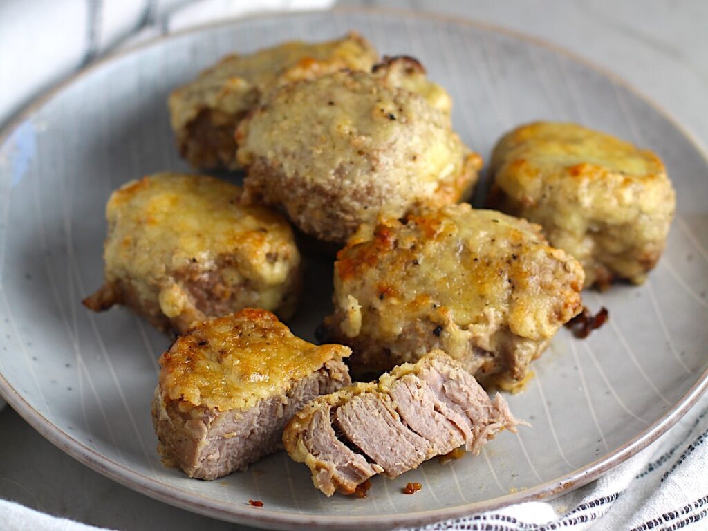 Parmesan Pork Tenderloin pieces on a plate with one cut in half. They are juicy, salty, and slightly cheesy. It's easy to make in the oven so you can enjoy it at home anytime!