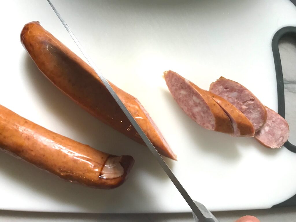 Knife slicing pre-cooked pork sausage on cutting board for Feijoada recipe.  It is rice and beans Brazilian style with pork sausages, beef, and more.
