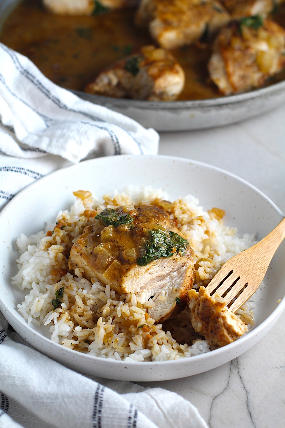 Fork holding a bit of a piece of Brazilian Chicken in Peanut Sauce on top of rice on a plate on counter with dish towel next to it. Skillet with chicken in background.