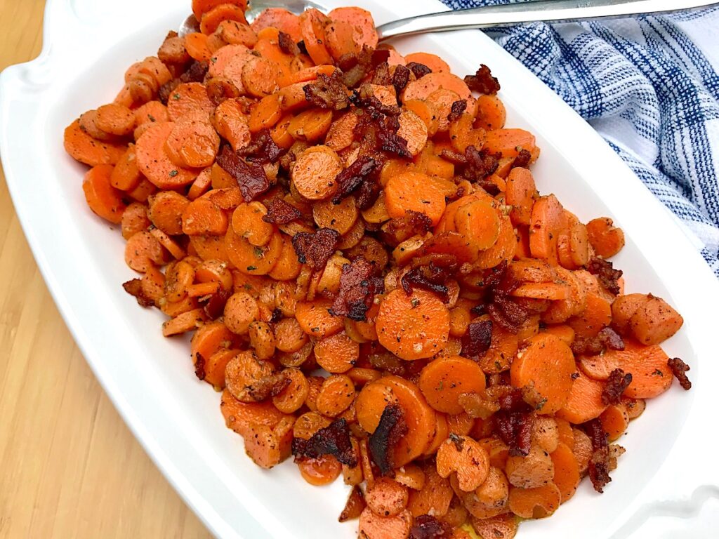 Carrot Bacon recipe on white serving platter with spoon in upper right corner and towel on table.