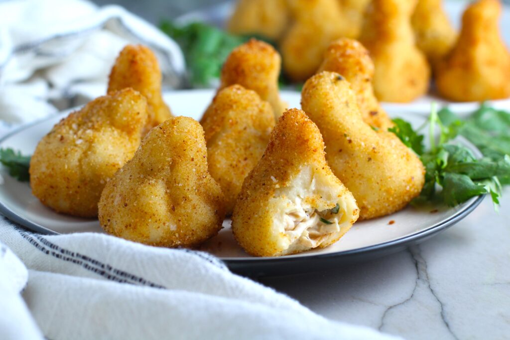 Golden brown breaded Brazilian Coxinha Chicken Croquettes on a plate with a bite taken out of the croquette in front. The plate has cilantro garnish and it's on counter with towel next to it.