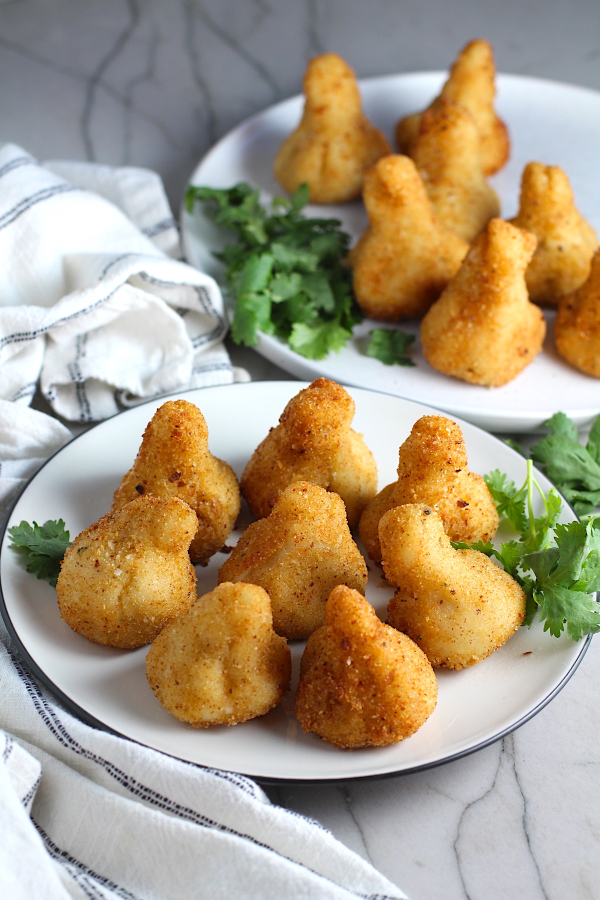 Golden brown breaded Brazilian Coxinha Chicken Croquettes on two plates with cilantro garnish on a counter with towel next to it.