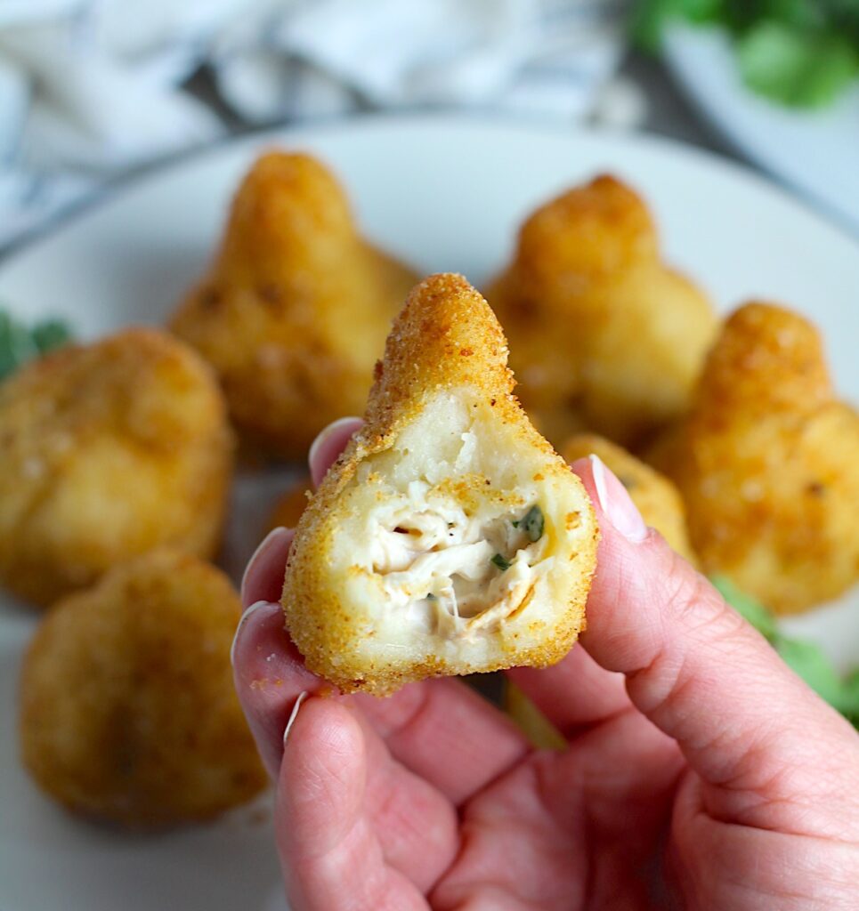 Golden brown breaded Brazilian Coxinha Chicken Croquettes on a plate with a hand holding one with a bite taken out of it. The plate has cilantro garnish and it's on counter with towel next to it.