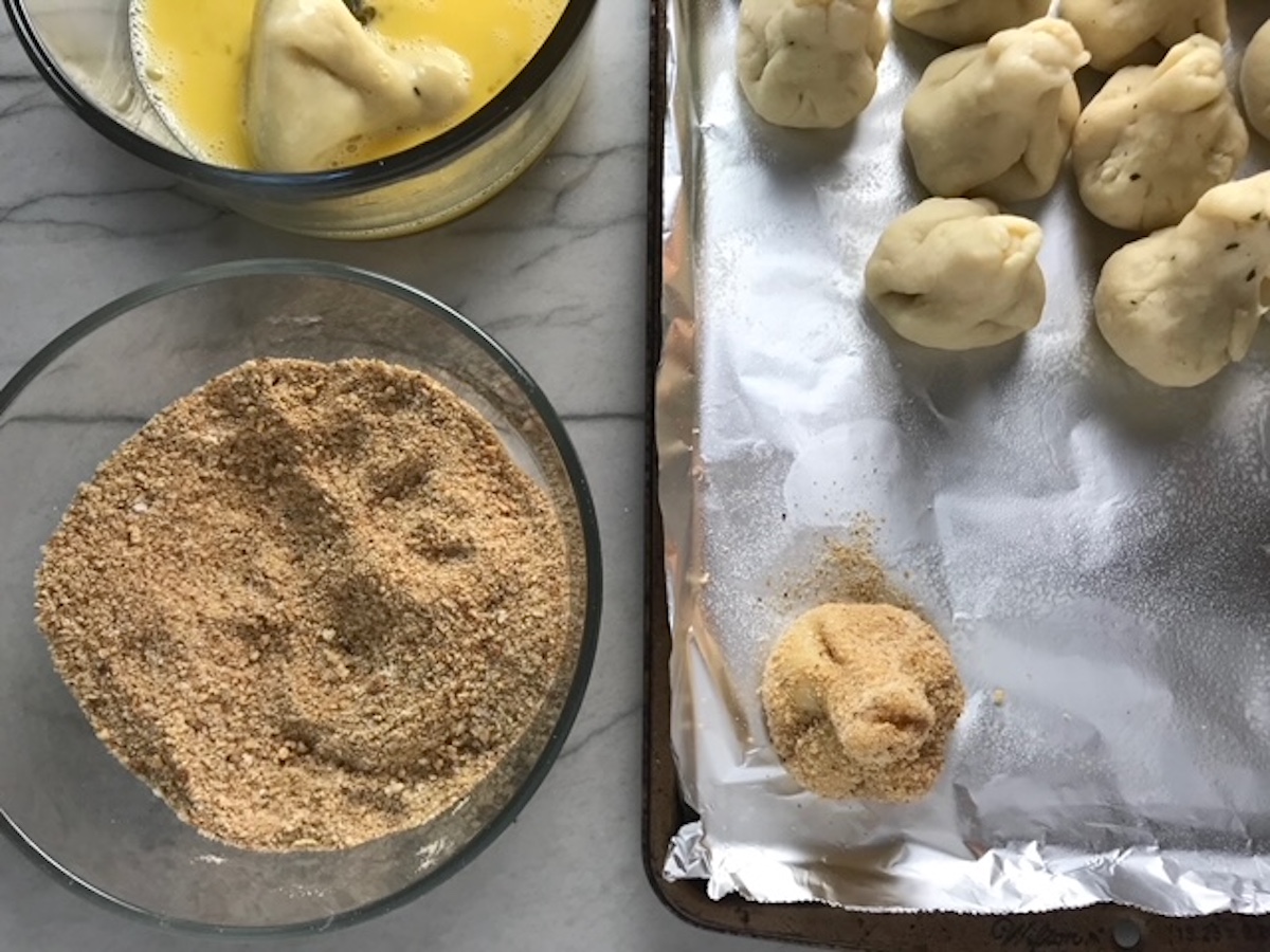 Bowl with beaten eggs, bowl with breadcrumbs, and sheet pan with one breaded croquette and others not yet breaded for Coxinha Chicken Croquettes.