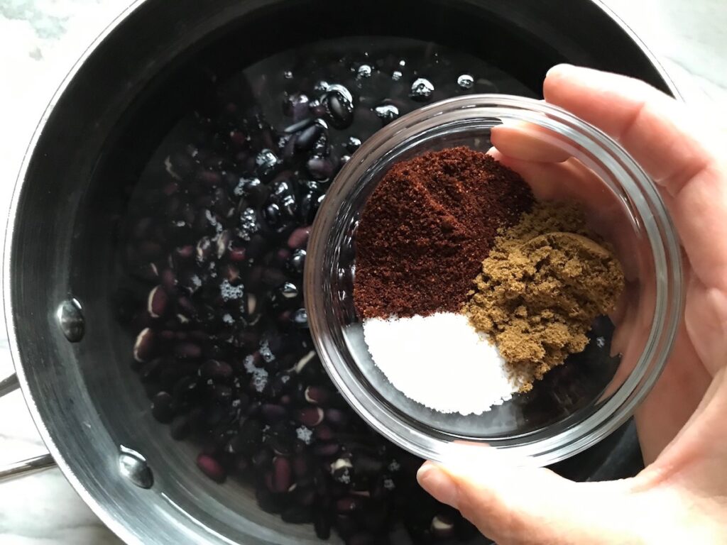 Hand about to pour seasoning over beans in water in a pot for black beans on counter for Brazilian Black Bean Puree, or Tutu de Feijão.