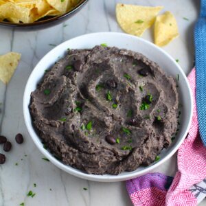 Brazilian Black Bean Puree in a bowl with cilantro pieces on top and tortilla chips on counter next to it. Known as Tutu de Feijao, it's a delicious side dish or topping to make ahead and have on hand!