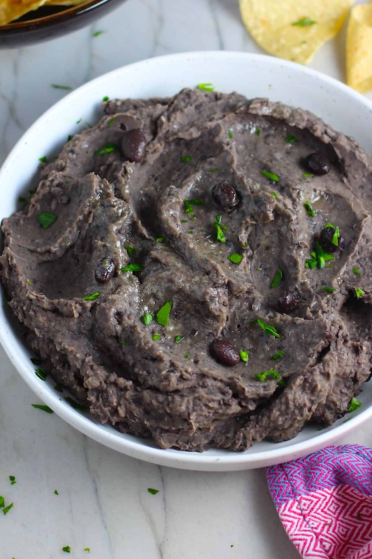 Brazilian Black Bean Puree in a bowl with cilantro pieces on top and tortilla chips on counter next to it.  Known as Tutu de Feijao, it's a delicious side dish or topping to make ahead and have on hand! 