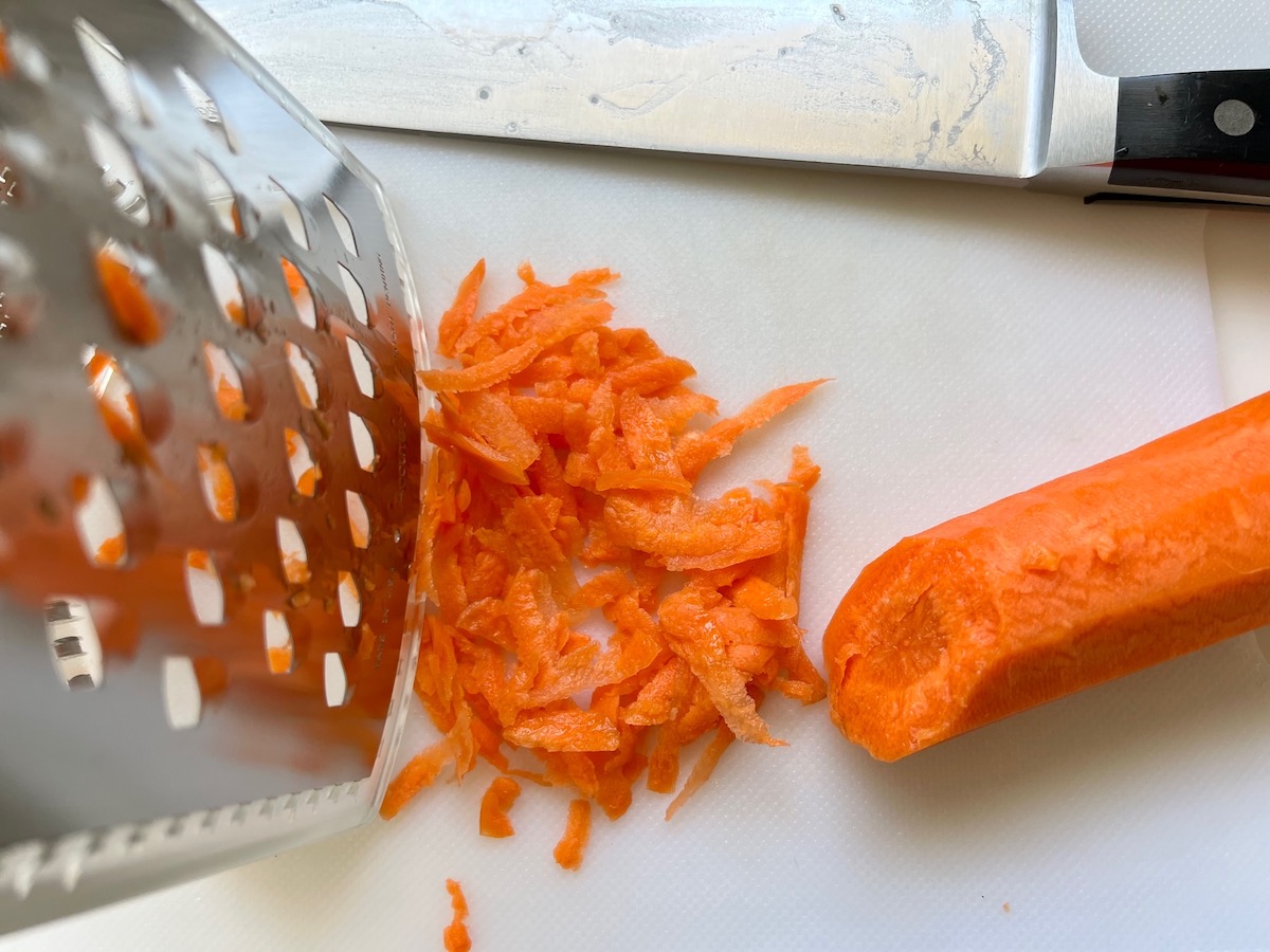 Whole carrot being shredded on a grater with shredded carrot sitting next to the carrot for Brazilian Potato Salad.