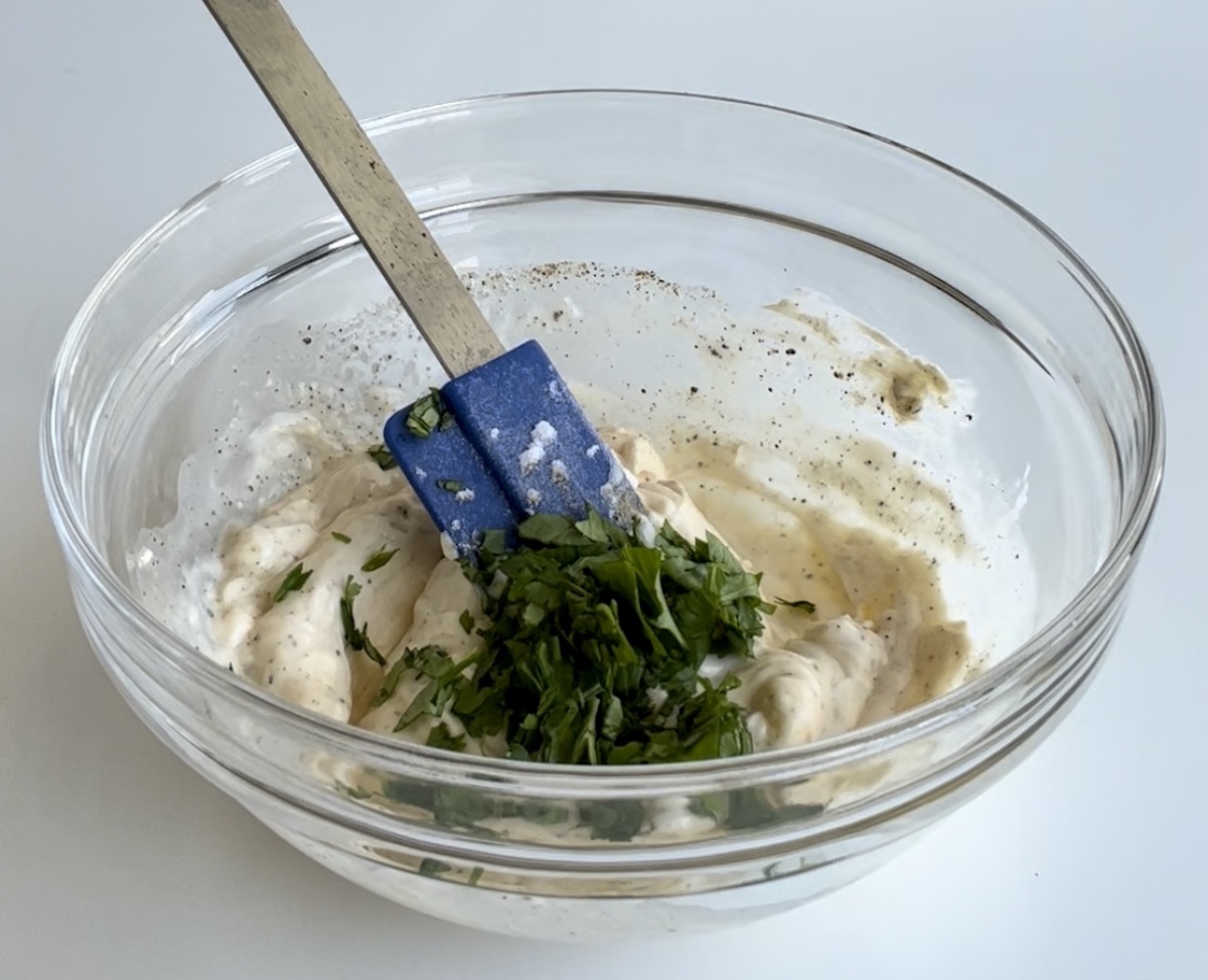 Brazilian Potato Salad dressing in a clear bowl with spatula and fresh cilantro and scallions added on top before being mixed in.