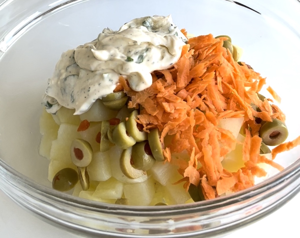 Cooked diced potatoes in a clear bowl with the sliced green olives with pimento and shredded carrots on top and the dressing sitting on top of everything before mixing it in for the Brazilian Potato Salad.