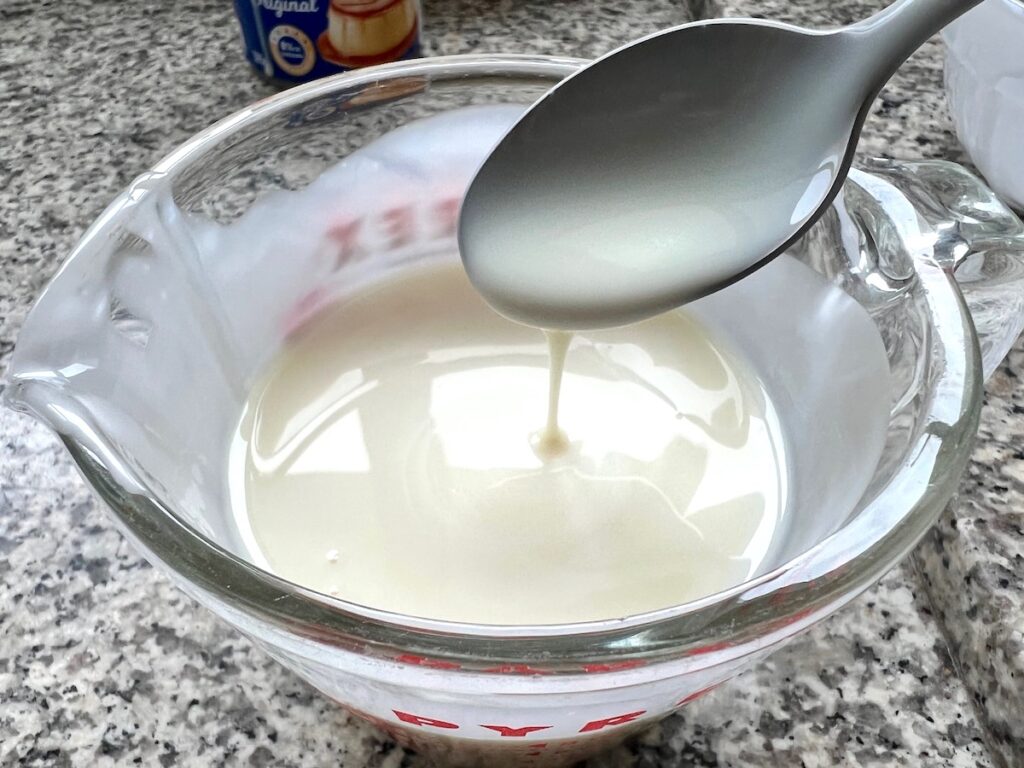 Glaze dripping from a spoon over a measuring cup of the glaze for Brazilian Coconut Cake.