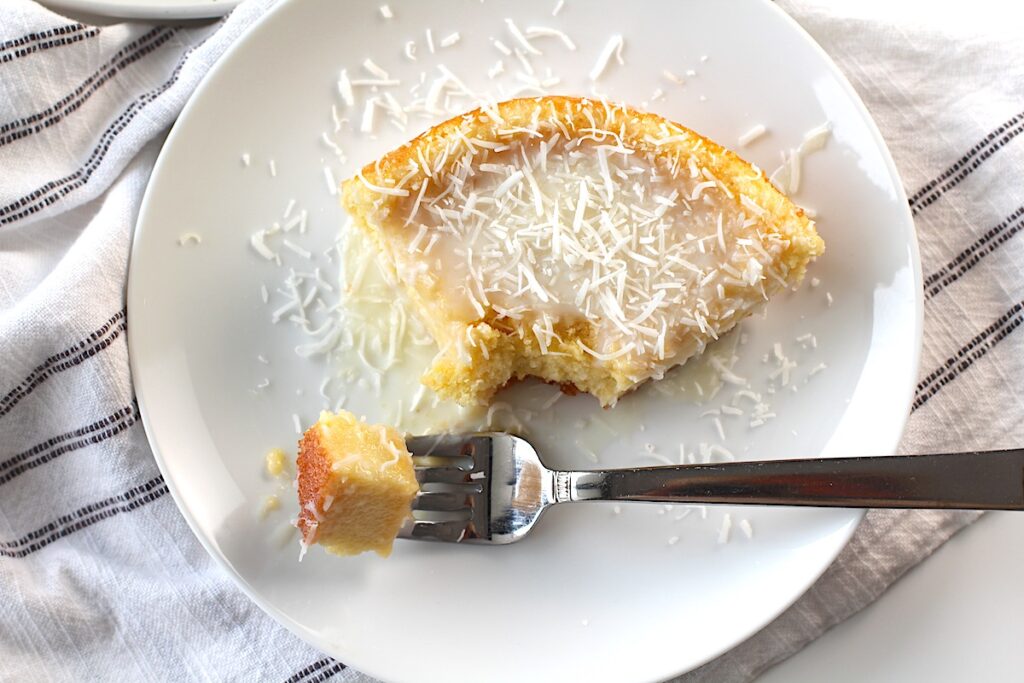 Brazilian Coconut Cake piece on a plate with a bite taken out and the rest of the cake on a platter in the background.