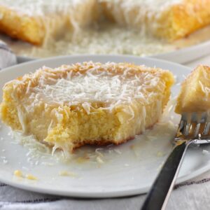 Brazilian Coconut Cake piece on a plate with a bite taken out and the rest of the cake on a platter in the background.
