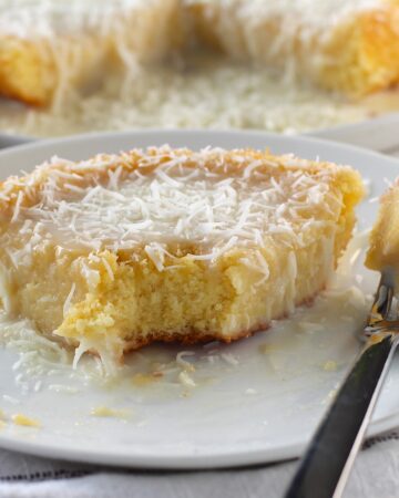 Brazilian Coconut Cake piece on a plate with a bite taken out and the rest of the cake on a platter in the background.