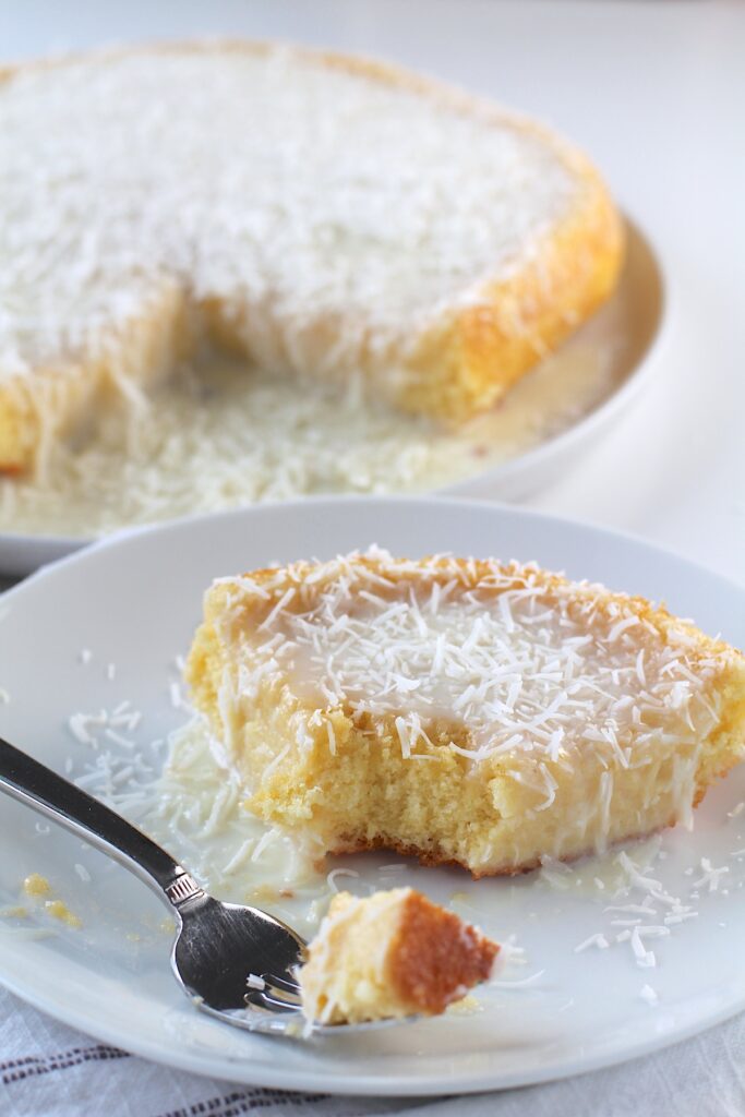 Brazilian Coconut Cake piece on a plate with a bite taken out and the rest of the cake on a platter in the background.