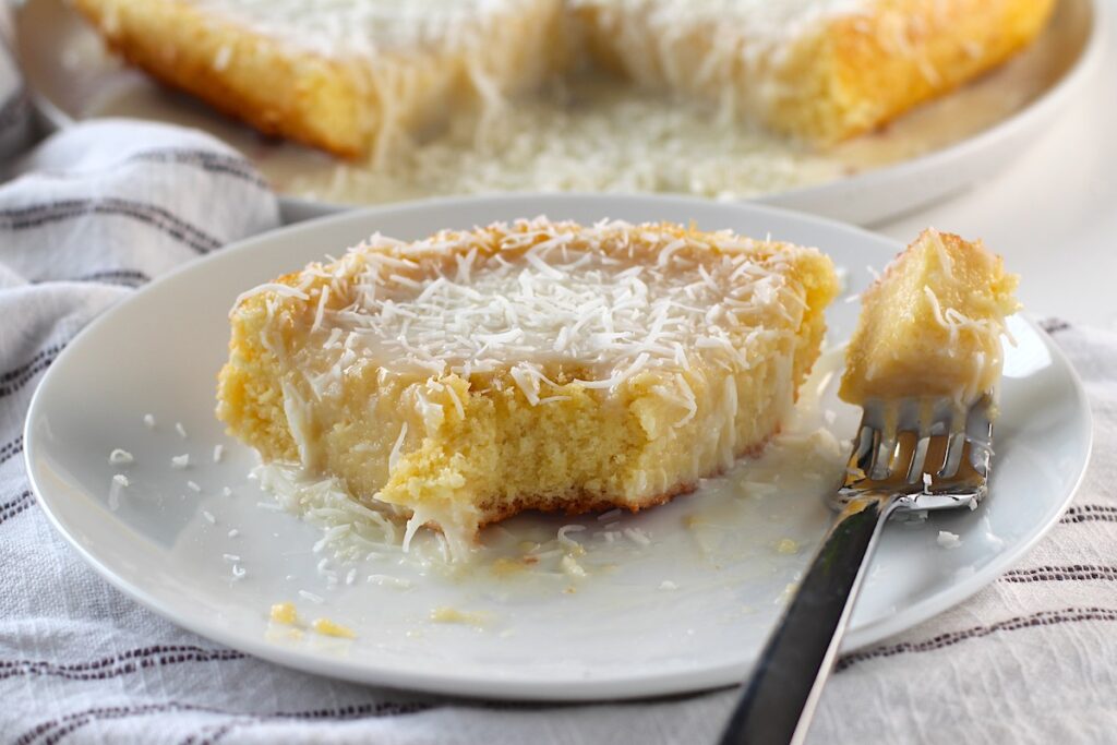 Brazilian Coconut Cake piece on a plate with a bite taken out and the rest of the cake on a platter in the background.