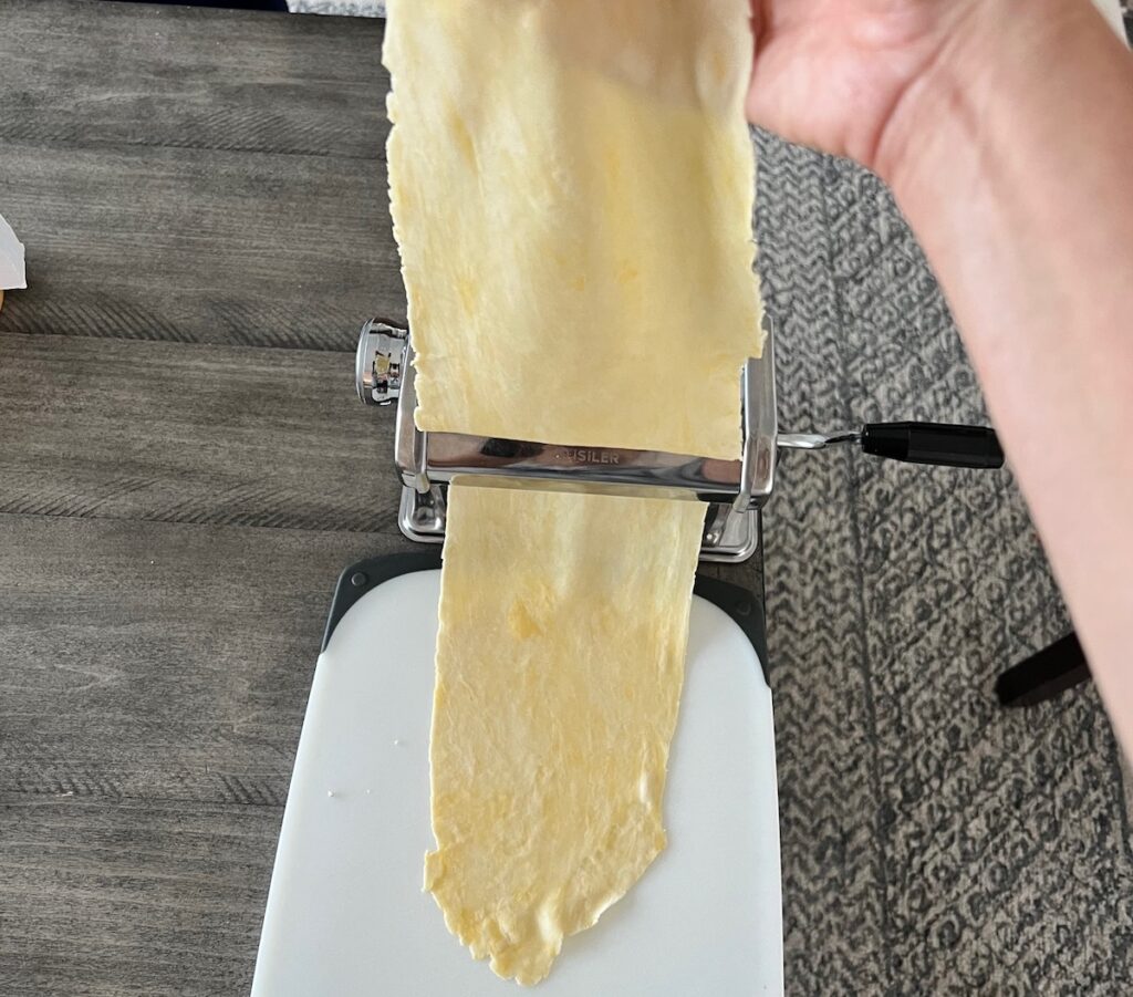 Hand holding thin dough sheet as it goes through the thinnest setting of the pasta rolling machine for Brazilian Pastel recipe.