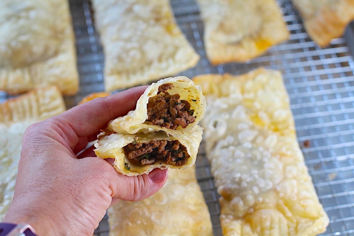Hand holding two halves stacked of golden brown fried Pastel over sheet pan of more pasteis for traditional Brazilian Pastel Recipe with ground beef filling.