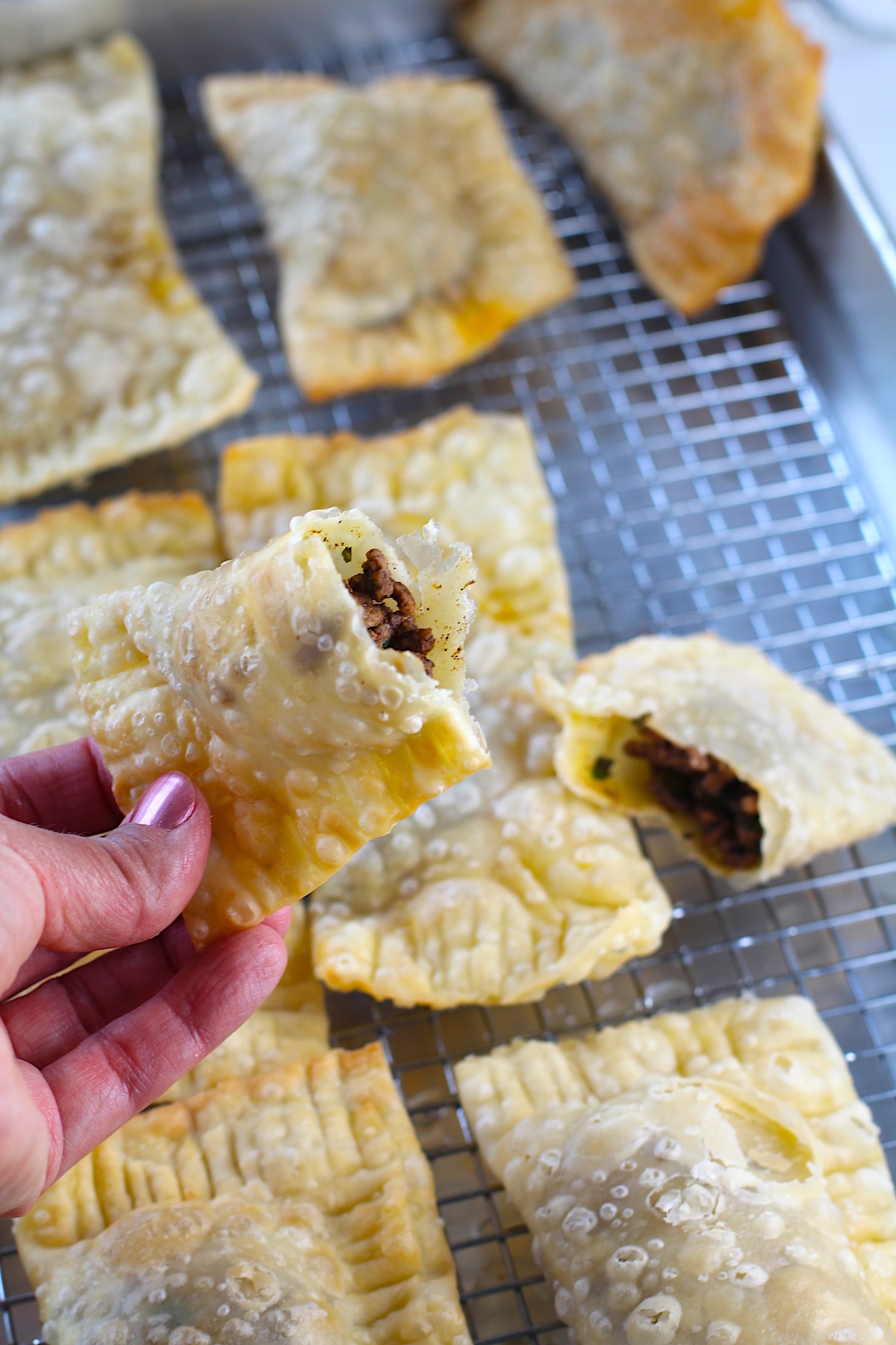 Hand holding half of golden brown fried Pastel over sheet pan of more pasteis for traditional Brazilian Pastel Recipe with ground beef filling.