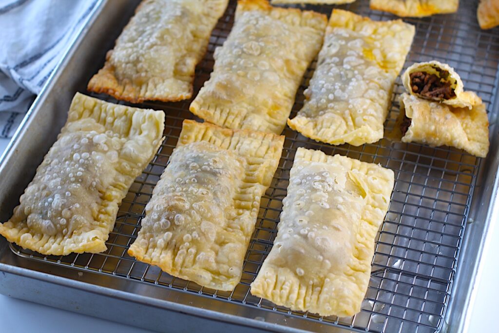 Six golden brown fried Pasteis on rack over sheet pan with another pastel cut open for traditional Brazilian Pastel Recipe with ground beef filling.
