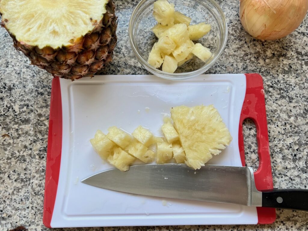 Pineapple being cut on a cutting board with a bowl of diced pineapple behind and half of the whole pineapple on the counter for Brazilian Marinated Chicken Stir Fry.