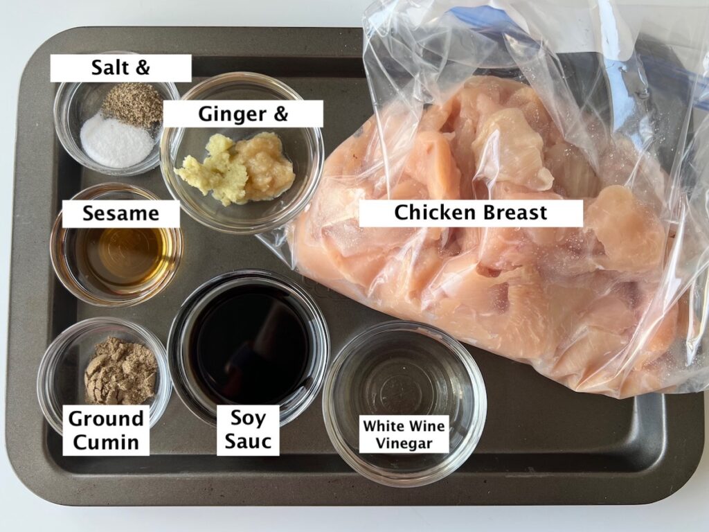 Ingredients prepped and measured out in small bowls for the chicken and marinade for Brazilian Marinated Chicken Stir Fry. From left top: salt and pepper, minced ginger and garlic, chicken breast strips, Sesame oil, ground cumin, soy sauce, white wine vinegar.
