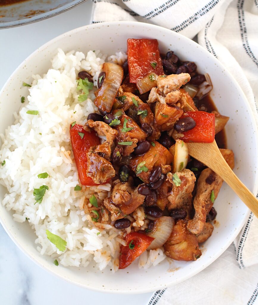 Brazilian Marinated Chicken Stir Fry with pineapple, black beans, red Pepper and onions in a bowl over white rice with a bamboo fork and cilantro garnish. It is truly one amazing dish with incredible flavor!