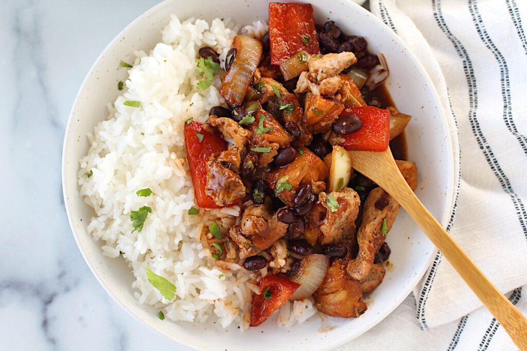 Brazilian Marinated Chicken Stir Fry with pineapple, black beans, red Pepper and onions in a bowl over white rice with a bamboo fork and cilantro garnish. It is truly one amazing dish with incredible flavor!